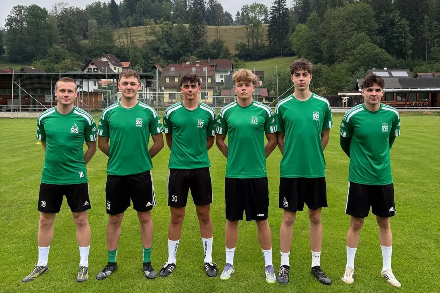 Sechs junge Fußballspieler in grünen Trikots stehen auf dem Rasen eines Fußballplatzes für ein Mannschaftsfoto. Im Hintergrund sind das Vereinsgelände, Wohnhäuser und bewaldete Hügel zu sehen.