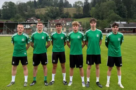 Acht Neuzugänge beim SV Stanz Sechs junge Fußballspieler in grünen Trikots stehen auf dem Rasen eines Fußballplatzes für ein Mannschaftsfoto. Im Hintergrund sind das Vereinsgelände, Wohnhäuser und bewaldete Hügel zu sehen.