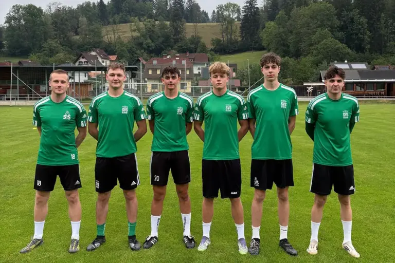 Sechs junge Fußballspieler in grünen Trikots stehen auf dem Rasen eines Fußballplatzes für ein Mannschaftsfoto. Im Hintergrund sind das Vereinsgelände, Wohnhäuser und bewaldete Hügel zu sehen.