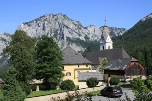 Blick auf die Pfarrkirche und den historischen Ortskern von Tragöß-St. Katharein vor der markanten Felskulisse. Zwischen Bäumen und traditionellen Gebäuden ragt der Kirchturm empor, im Vordergrund sind eine Straße und geparkte Autos zu sehen.