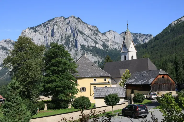 Pfarre Tragöß lädt zum Fest Blick auf die Pfarrkirche und den historischen Ortskern von Tragöß-St. Katharein vor der markanten Felskulisse. Zwischen Bäumen und traditionellen Gebäuden ragt der Kirchturm empor, im Vordergrund sind eine Straße und geparkte Autos zu sehen.