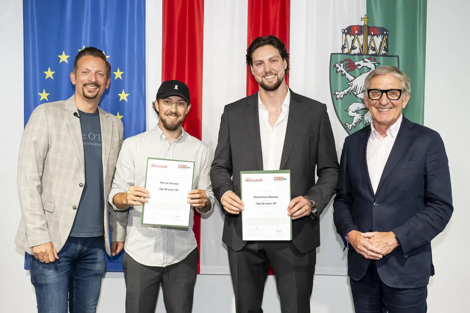 Vier Männer stehen vor den Flaggen der EU, Österreichs und der Steiermark. Zwei der Männer halten Urkunden der Auszeichnung „Top 30 unter 30“ in den Händen und lächeln in die Kamera.