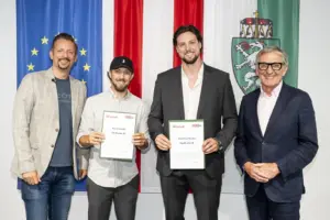 Vier Männer stehen vor den Flaggen der EU, Österreichs und der Steiermark. Zwei der Männer halten Urkunden der Auszeichnung „Top 30 unter 30“ in den Händen und lächeln in die Kamera.