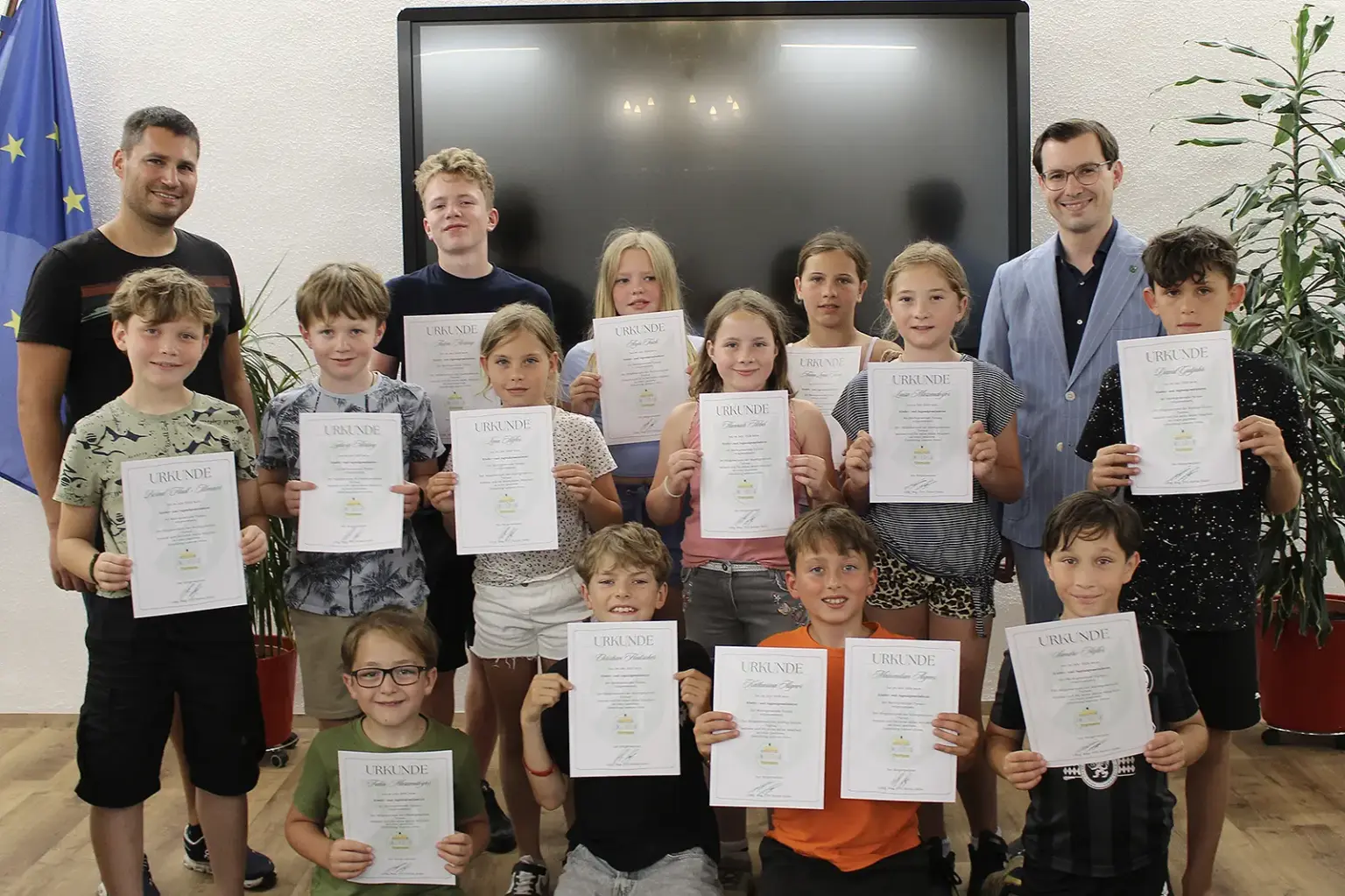 15 Kinder halten jeweils eine Urkunde in die Kamera. Hinter ihnen stehen zwei Männer. Im Hintergrund sind ein großer schwarzer Bildschirm, eine EU-Flagge sowie zwei Zimmerpflanzen zu sehen.