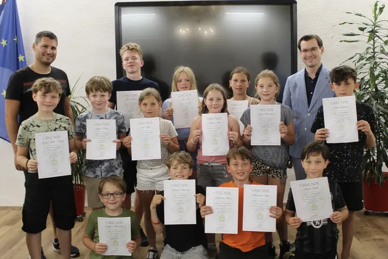 Turnauer Kindergemeinderat beendete seine Amtszeit 15 Kinder halten jeweils eine Urkunde in die Kamera. Hinter ihnen stehen zwei Männer. Im Hintergrund sind ein großer schwarzer Bildschirm, eine EU-Flagge sowie zwei Zimmerpflanzen zu sehen.