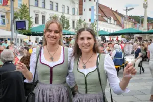 Zwei junge Frauen mit Trachtenkleidung und Krönchen sowie Weingläsern in einer Festgesellschaft auf einem innerstädtischen Platz.
