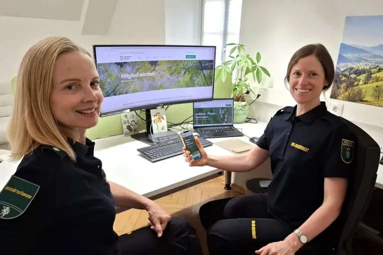 Berg- und Naturwacht mit neuem Webauftritt Zwei Frauen sitzen lächelnd an einem Schreibtisch mit Breitbild-Computermonitor