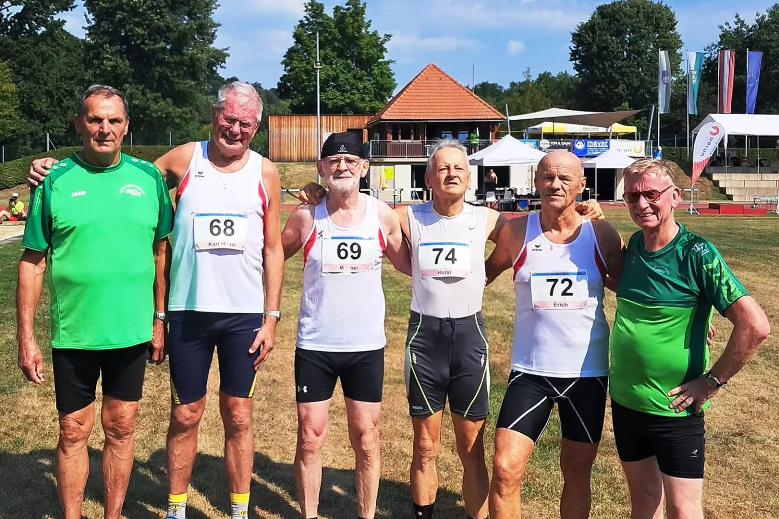 Sechs Männer in Sportbekleidung stehen auf einer Leichtathletikanlage nebeneinander und posieren für ein Gruppenfoto. Vier von ihnen tragen weiße Laufshirts mit Startnummern (68, 69, 72 und 74), zwei grüne Vereinsshirts. Im Hintergrund sind Tribünen, Zelte, Fahnen und ein Gebäude mit rotem Ziegeldach zu sehen.