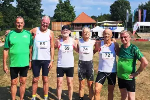 Sechs Männer in Sportbekleidung stehen auf einer Leichtathletikanlage nebeneinander und posieren für ein Gruppenfoto. Vier von ihnen tragen weiße Laufshirts mit Startnummern (68, 69, 72 und 74), zwei grüne Vereinsshirts. Im Hintergrund sind Tribünen, Zelte, Fahnen und ein Gebäude mit rotem Ziegeldach zu sehen.