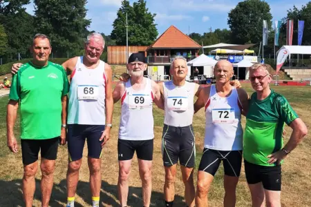 Sechs Männer in Sportbekleidung stehen auf einer Leichtathletikanlage nebeneinander und posieren für ein Gruppenfoto. Vier von ihnen tragen weiße Laufshirts mit Startnummern (68, 69, 72 und 74), zwei grüne Vereinsshirts. Im Hintergrund sind Tribünen, Zelte, Fahnen und ein Gebäude mit rotem Ziegeldach zu sehen.