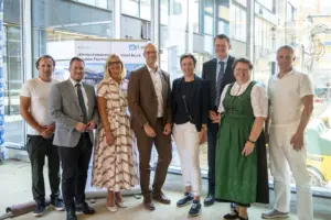 Acht Personen stehen nebeneinander in einem hellen Gebäude auf einer Baustelle. Hinter ihnen befindet sich ein Aufsteller mit der Aufschrift „LKH Hochsteiermark Standort Bruck – Neubau Psychiatrie“. Durch die großen Glasflächen sind weitere Gebäudeteile und Baustelleneinrichtungen zu sehen.