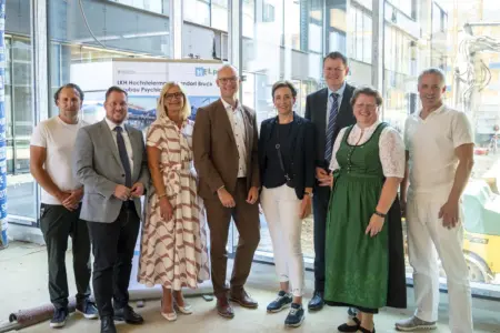 LKH Hochsteiermark: Gleichenfeier für Psychiatrie-Abteilung in Bruck Acht Personen stehen nebeneinander in einem hellen Gebäude auf einer Baustelle. Hinter ihnen befindet sich ein Aufsteller mit der Aufschrift „LKH Hochsteiermark Standort Bruck – Neubau Psychiatrie“. Durch die großen Glasflächen sind weitere Gebäudeteile und Baustelleneinrichtungen zu sehen.