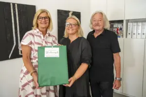 Zwei Frauen und ein Mann stehen in einem Büro vor zwei schwarzen Wandbildern. Die beiden Frauen halten gemeinsam ein großes, grün verpacktes Geschenk mit einem Anhänger, auf dem „herzlichen Glückwunsch“ steht. Im Hintergrund befinden sich weiße Schränke mit Aktenordnern.
