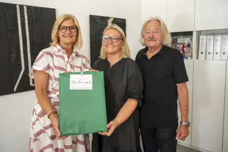 Zwei Frauen und ein Mann stehen in einem Büro vor zwei schwarzen Wandbildern. Die beiden Frauen halten gemeinsam ein großes, grün verpacktes Geschenk mit einem Anhänger, auf dem „herzlichen Glückwunsch“ steht. Im Hintergrund befinden sich weiße Schränke mit Aktenordnern.