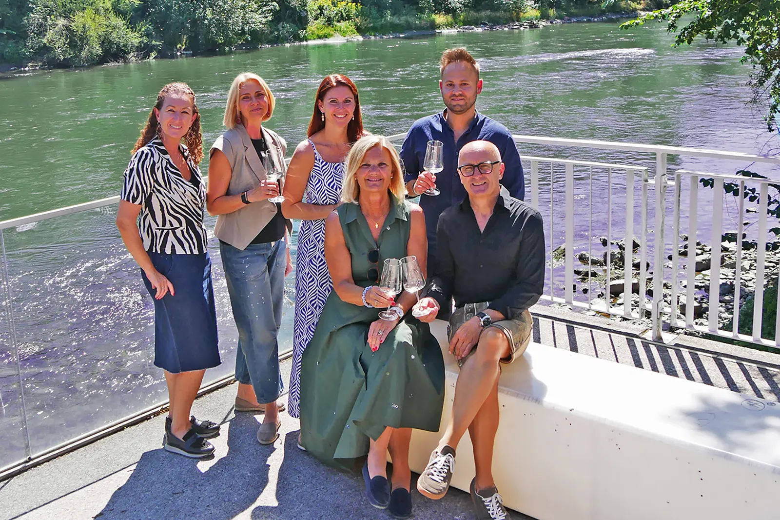 Zwei Männer und vier Frauen stehen und sitzen mit Weingläsern an einem Flussufer