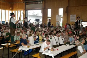 Eine Festhalle mit Publikum auf Biertischgarnituren. Vier Männer stehen auf Tischen und schwingen Peitschen.
