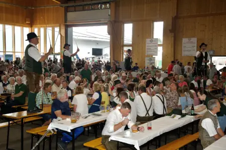 Eine Festhalle mit Publikum auf Biertischgarnituren. Vier Männer stehen auf Tischen und schwingen Peitschen.