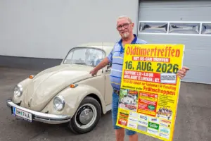 Ein Mann hält ein grelles Plakat in einer Hand, mit der anderen zeigt er auf ein Auto des Fabrikats VW Käfer