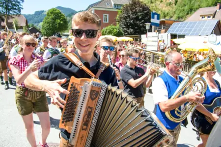 Tausende Gäste beim Musi-Kirtag in Gasen Ein junger Mann mit Sonnenbrille und steirischer Harmonika auf einem Dorfplatz, im Hintergrund zahlreiche weitere Musiker, großteils mit Blasinstrumenten