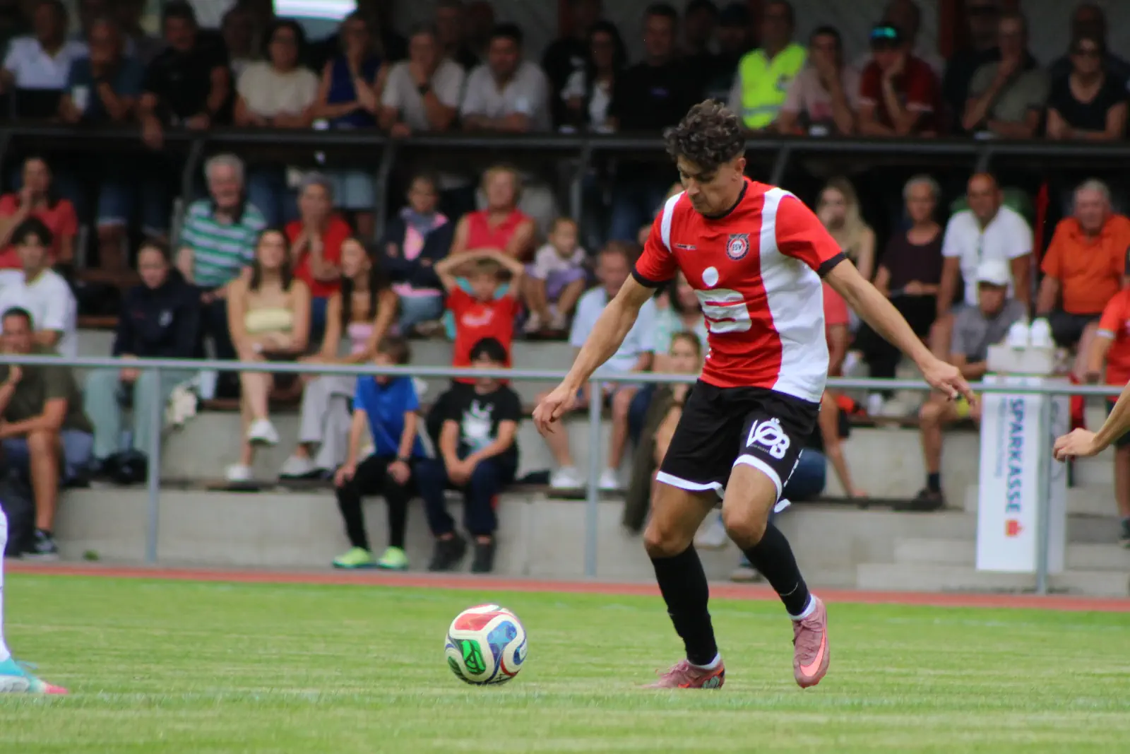 Ein Fußballer mit einem Ball, dahinter sitzende Zuseher.
