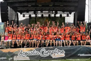 Rund 80 Menschen in orangen Shirts winkend und lachend auf einer Open-Air-Bühne