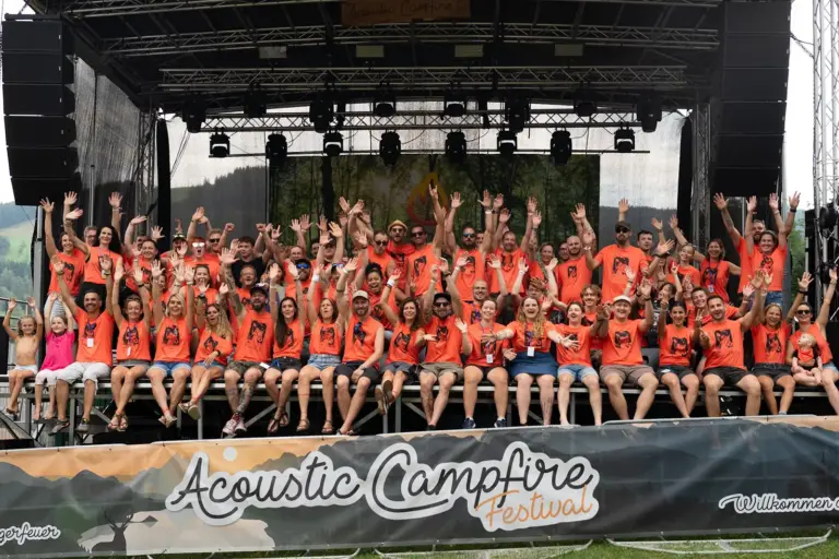 Rund 80 Menschen in orangen Shirts winkend und lachend auf einer Open-Air-Bühne