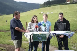 Vier Personen stehen bei einer Pressekonferenz auf einer Wiese vor mehreren Mikrofonen. Ein Schild am Tisch warnt davor, dass Hundekot und Müll im Futter Tiere gefährden.