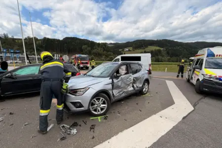 Ein Pkw mit Beschädigung an der Fahrerseite auf einer Straße, ein Feuerwehrmann steht am Fahrzeug, im Hintergrund ein weiterer Pkw, ein Lieferwagen und je ein Einsatzfahrzeug der Feuerwehr und des Roten Kreuzes