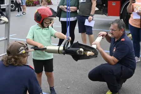 Ein Kind mit Helm schneidet mit einer Bergeschere ein Stück Holz durch, das von einem Feuerwehrmann gehalten wird