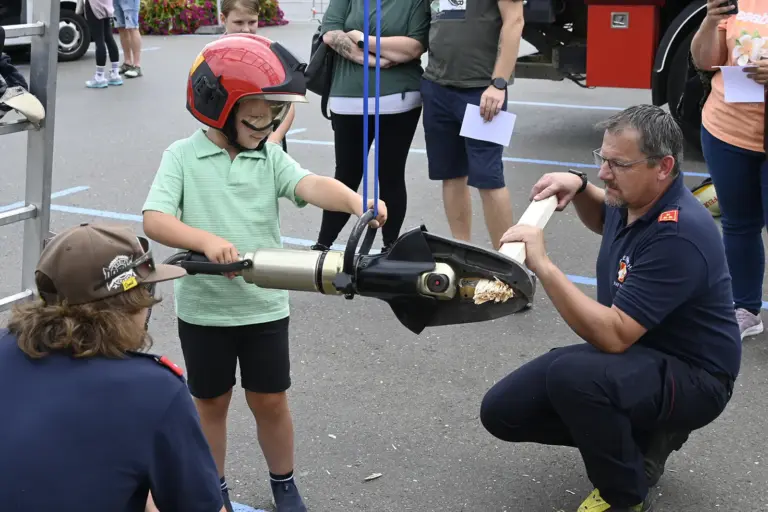 Ein Tag mit den Trofaiacher Einsatzkräften Ein Kind mit Helm schneidet mit einer Bergeschere ein Stück Holz durch, das von einem Feuerwehrmann gehalten wird