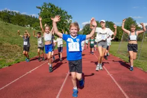 Mehrere Kinder mit Startnummern laufen auf einer Tartanbahn auf die Kamera zu und haben dabei die Arme erhoben. Im Vordergrund trägt ein Kind im blauen Trikot die Startnummer 11. Im Hintergrund sind weitere Kinder auf der Laufbahn, eine Wiese, Bäume und ein Fußballtor zu sehen.