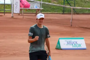 Ein junger Tennisspieler steht mit Tennisschläger auf einem Sandplatz und hält eine geballte Faust hoch. Er trägt ein dunkles Sportshirt und eine weiße Kappe. Im Hintergrund sind das Tennisnetz und eine Werbetafel mit der Aufschrift „Die Stadt Bruck an der Mur“ zu sehen.
