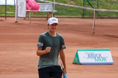 Ein junger Tennisspieler steht mit Tennisschläger auf einem Sandplatz und hält eine geballte Faust hoch. Er trägt ein dunkles Sportshirt und eine weiße Kappe. Im Hintergrund sind das Tennisnetz und eine Werbetafel mit der Aufschrift „Die Stadt Bruck an der Mur“ zu sehen.
