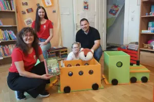 Zwei Frauen und ein Mann hocken neben einem Holzzug mit einem Kind darin