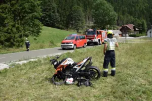 Ein beschädigtes Motorrad in einer Wiese, daneben ein Feuerwehrmann, im Hintergrund zwei Feuerwehrfahrzeuge auf einer Straße