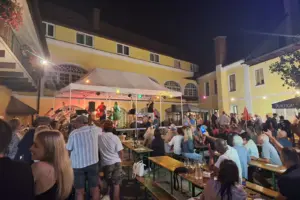 Zehntes Sommerkonzert in Ratten Der Innenhof eines Hauses mit gelber Fassade, eine Bühne mit Zeltdach ist aufgebaut, darauf eine Band, im Vordergrund Publikum