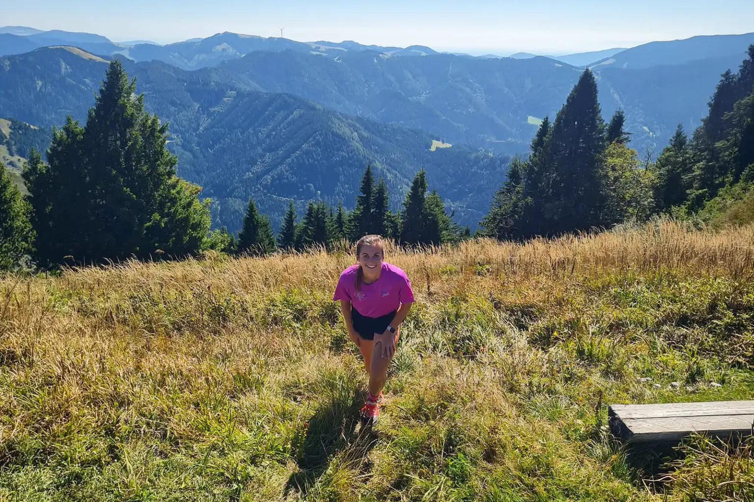 Stanzer Trailrun: 48 Kilometer im Laufschritt Eine Läuferin in pinkem Shirt steht auf einer sonnigen Bergwiese. Im Hintergrund erstreckt sich eine bewaldete Berglandschaft.