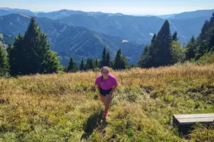 Eine Läuferin in pinkem Shirt steht auf einer sonnigen Bergwiese. Im Hintergrund erstreckt sich eine bewaldete Berglandschaft.