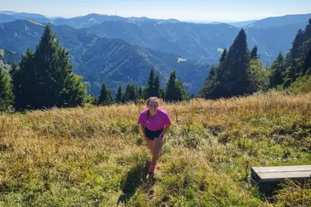Eine Läuferin in pinkem Shirt steht auf einer sonnigen Bergwiese. Im Hintergrund erstreckt sich eine bewaldete Berglandschaft.