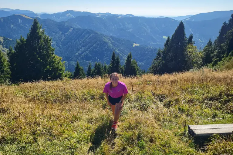 Stanzer Trailrun: 48 Kilometer im Laufschritt Eine Läuferin in pinkem Shirt steht auf einer sonnigen Bergwiese. Im Hintergrund erstreckt sich eine bewaldete Berglandschaft.