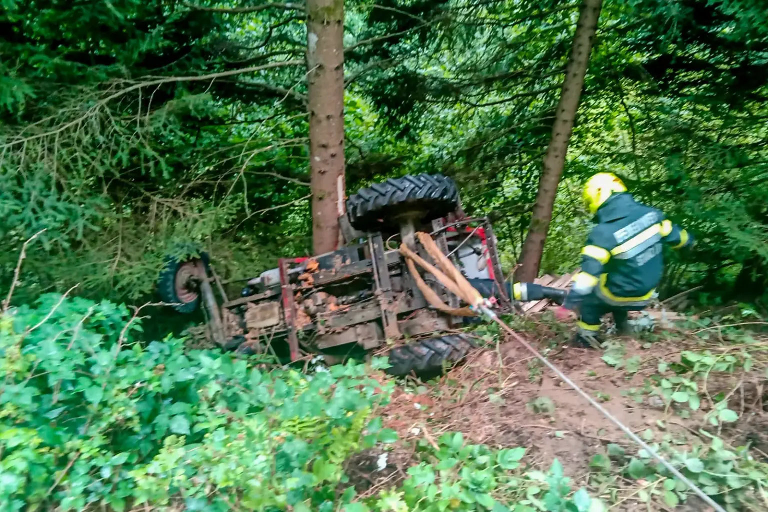 Ein Traktor liegt seitlich in einem Waldstück, an der Achse sind ein Bergungsgurt und ein Seil befestigt, ein Feuerwehrmann nähert sich dem Fahrzeug