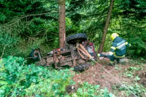 Ein Traktor liegt seitlich in einem Waldstück, an der Achse sind ein Bergungsgurt und ein Seil befestigt, ein Feuerwehrmann nähert sich dem Fahrzeug