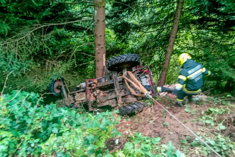 Traktorabsturz in Stanz, Fahrer unverletzt Ein Traktor liegt seitlich in einem Waldstück, an der Achse sind ein Bergungsgurt und ein Seil befestigt, ein Feuerwehrmann nähert sich dem Fahrzeug