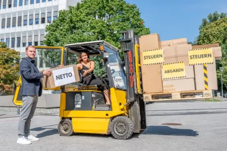 Heute ist „Tax Freedom Day“ Eine Frau sitzt auf einem mit einer Palette voll Schachteln beladenen Gabelstapler, neben dem Fahrzeug steht ein Mann mit einer Schachtel in Händen.