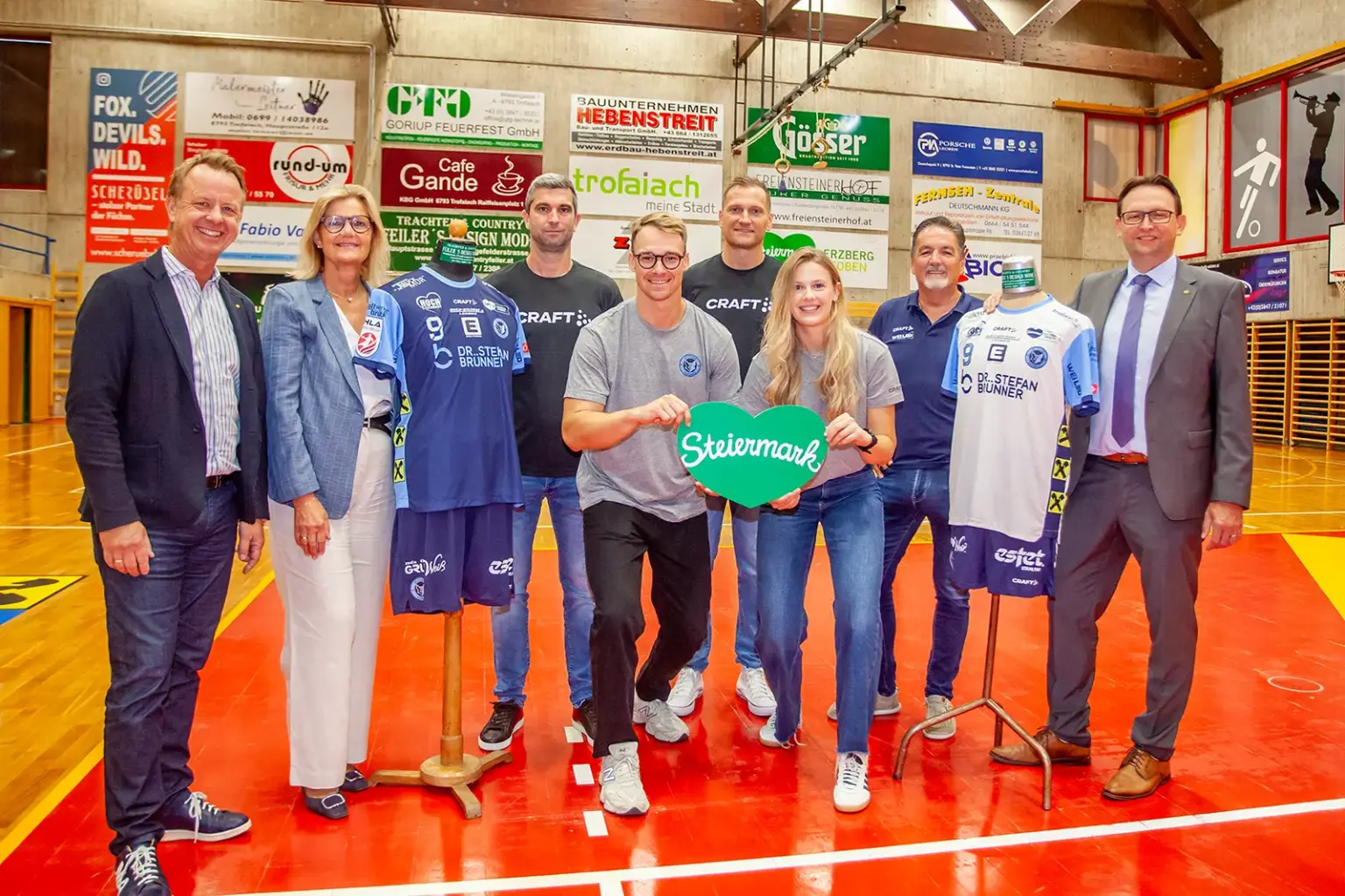 Bruck-Trofaiach: Mit Obersteirer-Power in die neue Handballsaison Zwei Frauen, sechs Männer und zwei Sportkleidung tragende Kleiderpuppen in einer Sporthalle vor einer Wand voller Werbetafeln.