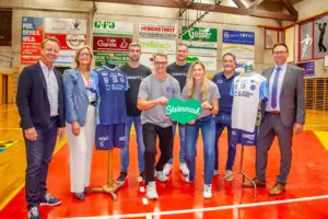 Zwei Frauen, sechs Männer und zwei Sportkleidung tragende Kleiderpuppen in einer Sporthalle vor einer Wand voller Werbetafeln.