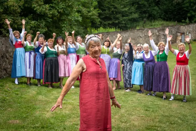 Trofaiach: Die Musik ist ihr halbes Leben Eine Frau mit Blumenkranz im grauen Haar steht auf einer Wiese, im Hintergrund 15 Frauen in Trachtenkleidung, die Hände jubelnd in die Höhe gestreckt