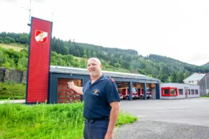 Ein Mann in Feuerwehrkleidung steht vor einem grau-roten Rüsthaus mit rotem Turm