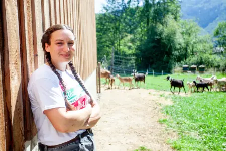In Eigenregie zum Traumberuf Bäuerin Eine Frau mit geflochtenen Zöpfen steht mit verschränkten Armen an eine Holzwand gelehnt, im Hintergrund eine Ziegenherde