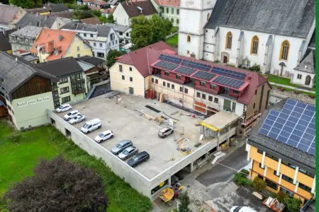 Die Neugestaltung des Aflenzer Ortszentrums ist im Finale Luftaufnahme eines Gebäudekomplexes mit roten Dächern und Photovoltaikanlagen. Vor dem teilweise eingerüsteten Gebäude befindet sich eine große Betonfläche mit parkenden Autos und Baumaterialien.